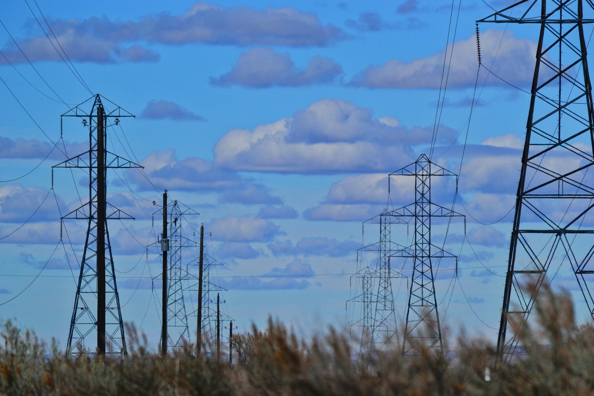 Transmission towers and power lines silhouetted against a bright blue sky with clouds.